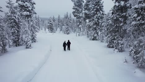 Drone-Follows-Couple-Walking-Together-Exploring-In-Snowy-Forest-in-Lapland,-Finland,-Arctic-Circle
