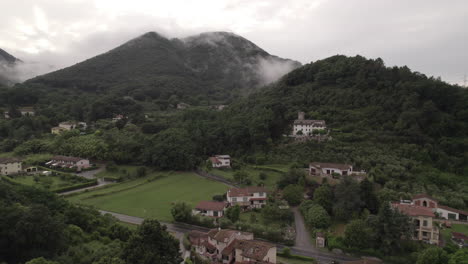Drone-shot-of-clouds-moving-past-the-camera-in-the-hills-and-mountains-in-Italy-on-a-cloudy-grey-day-with-villages-and-houses-down-on-the-ground-in-between-green-fields-LOG