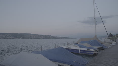 Reiseaufnahme-Auf-Einem-Holzsteg-Mit-Blick-Auf-Den-Zürichsee,-Mit-Booten-Im-Vordergrund-Während-Des-Sonnenuntergangs-In-Zeitlupe-Und-Bergen-Im-Hintergrund-Und-Wellen-Im-Stausee