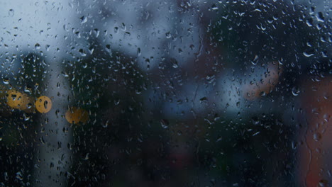 Regentropfen-Rieseln-An-Einem-Fenster-Herunter,-Im-Hintergrund-Sind-Unscharfe-Lichter-Zu-Sehen