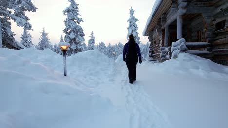 Girl-Walks-Alongside-Cabin-in-Snowy-Winter-Wonderland-in-Lapland,-Finland,-Arctic-Circle