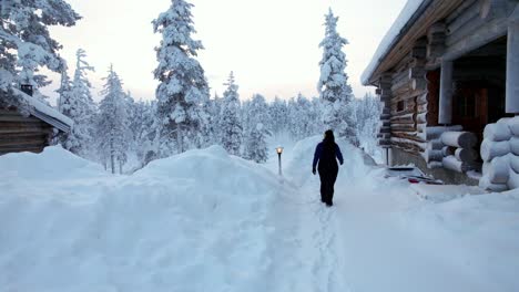 Girl-Exploring-Alongside-Cabin-in-Snowy-Winter-Wonderland-In-Lapland,-Finland,-Arctic-Circle