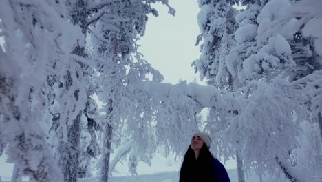 Girl-Looking-Up-Under-Trees-In-Snowy-Forest-In-Snowy-Winter-Wonderland-In-Lapland,-Finland,-Arctic-Circle