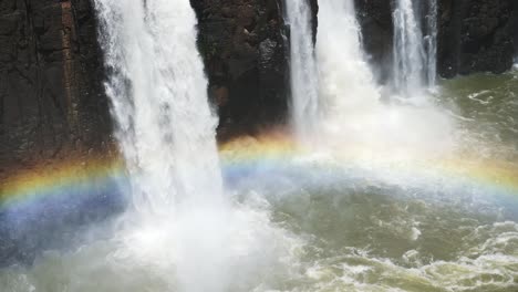 Bright-Beautiful-Rainbows-Covered-by-Huge-Slow-Motion-Waterfalls-in-Iguacu-Falls,-Water-Falling-of-Dark-Rocky-Cliff-Edge-onto-Colourful-Rainbow-Arch-in-Iguazu-Falls,-Brazil