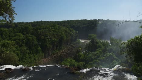 Iguazu-Falls-Waterfall-in-Argentina,-Slo-Mo-Waterfall-Stream-Falling-off-Huge-Cliff-into-Distant-Jungle-Rainforest-Landscape,-Tall-Greenery-Surrounding-Beautiful-Clear-Water-Dropping-off-Cliff-Edge