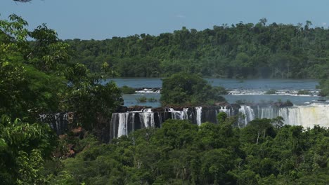 Distant-Waterfalls-over-Tall-Wild-Jungle-Trees,-Big-Bright-Green-Dense-Treelife-Surrounding-Large-Body-of-Water-Pouring-Down-Steep-Ciff-Edge,-Wide-Long-Waterfall-in-Iguazu-Falls,-Brazil