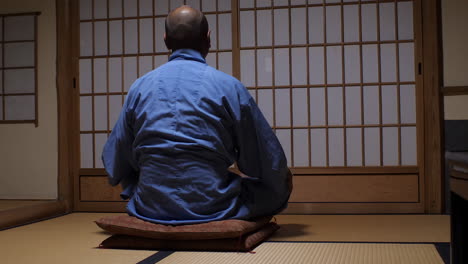 Hombre-Con-Traje-Tradicional-Sentado-Serenamente-En-Una-Habitación-Japonesa,-Suelo-De-Tatami-Y-Puertas-Shoji-Al-Fondo.