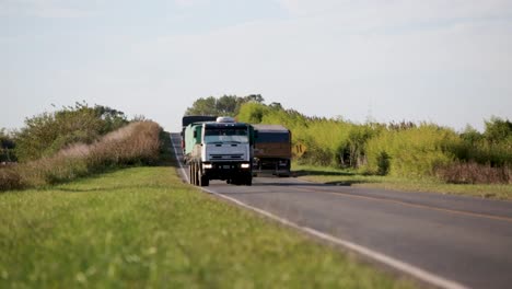 Convoys-of-trucks-driving-on-a-rural-road-with-lush-greenery,-sunny-day