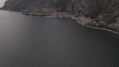 Droneshot-Sobre-La-Ciudad-Limone-Italia-En-El-Lago-De-Garda-En-Un-Día-Gris-Con-Montañas,-Nubes-Y-Troncos-De-Agua
