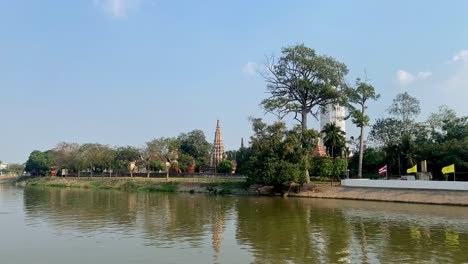 Tranquil-river-view-with-traditional-Thai-temple-pagoda,-lush-greenery,-and-clear-skies