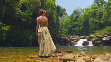 Woman-in-a-dress-standing-by-a-tropical-waterfall,-sunlight-filtering-through-the-forest,-serene