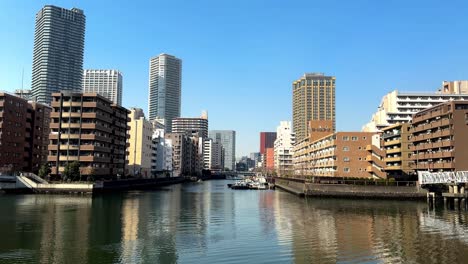 Sunlit-cityscape-with-modern-buildings-along-a-calm-river,-clear-blue-sky,-urban-waterfront-scene