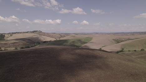 Toma-De-Drones-De-Caminos-Sinuosos-Y-Campos-Agrícolas-Dorados-En-El-Paisaje-De-Toscana-Italia-En-Un-Día-Soleado-Con-Cielo-Azul-Y-Nubes-En-El-Horizonte-Proyectando-Un-Tronco-De-Sombra-En-Movimiento