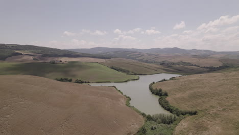 Drone-shot-of-winding-roads-and-golden-farm-fields-in-Tuscany-Italy-landscape-on-a-sunny-day-with-blue-sky-and-clouds-on-the-horizon-and-a-small-lake-or-puddle-in-the-middle-of-the-scene-LOG
