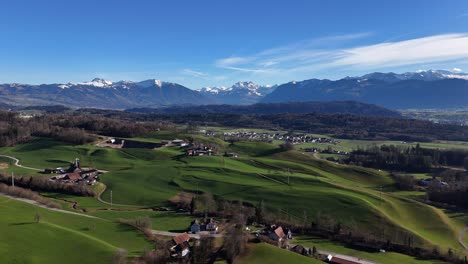 Los-Alpes-Glarner-Siguen-Al-Pueblo-De-Jona-En-Suiza,-Exuberantes-Campos-Verdes-Bajo-Un-Cielo-Azul-Claro,-Vista-Aérea