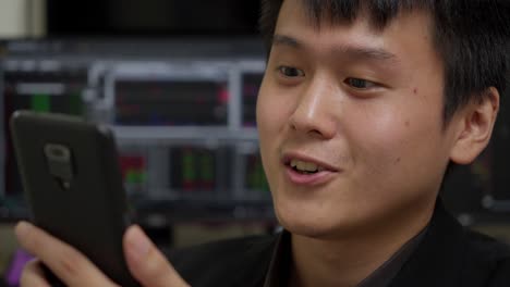 Junger-Asiatischer-Mann-Lächelt,-Während-Er-Auf-Sein-Smartphone-Schaut,-Büroumgebung-Mit-Börsendiagrammen-Im-Hintergrund