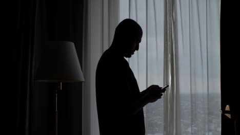 Silhouette-of-bald-man-standing-near-the-window-with-the-curtains-closed,-typing-on-a-mobile-phone-screen-Static-Shot