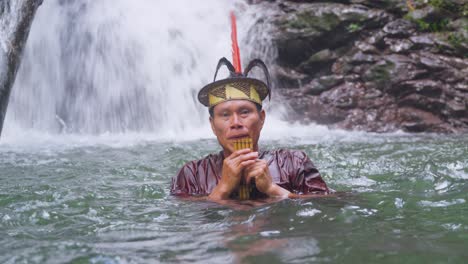 Man-in-traditional-attire-emerges-from-water-at-waterfall-in-pucallpa,-Peru-playing-flute