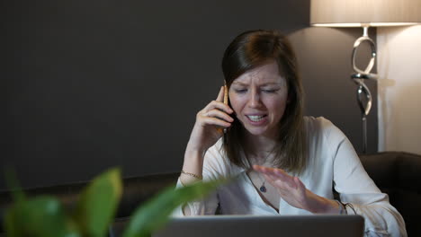 Angry-young-professional-woman-talking-on-the-phone-frustrated-working-at-home-in-front-of-a-laptop-computer
