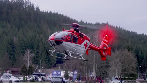 Red-and-white-rescue-helicopter-taking-off-from-an-open-field-with-parked-cars-and-forest-in-the-background
