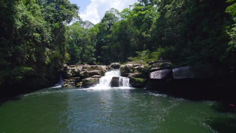 Lush-greenery-surrounds-a-serene-waterfall-in-Oxapampa,-Peru,-with-a-native-person-observing-from-above,-daylight
