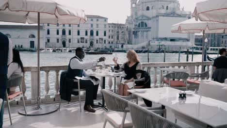 Ein-Edel-Gekleidetes-Paar-Mit-Sonnenbrille-Jubelt-Seinen-Champagnergläsern-Zu,-Isst-Eis-Und-Genießt-Die-Aussicht-Auf-Einer-Terrasse-Direkt-Am-Canal-Grande-In-Venedig,-Italien-–-Luxus-Jetset-Reiselebensstil