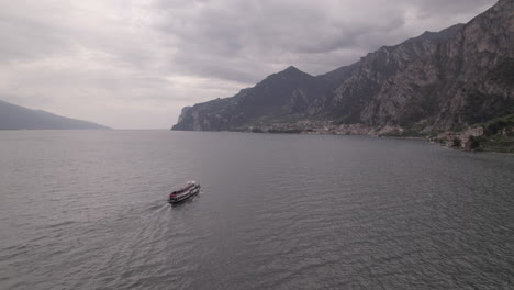 Droneshot-of-a-ferry-boat-going-to-the-city-Limone-Italy-on-Lake-Garda-on-a-grey-day-with-mountains-and-water-LOG