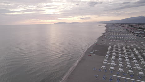 Drone-shot-above-the-beaches-in-Viareggio-Italy-during-sunset-on-a-grey-day-flying-towards-the-sea-with-buildings-and-mountains-in-the-background-LOG