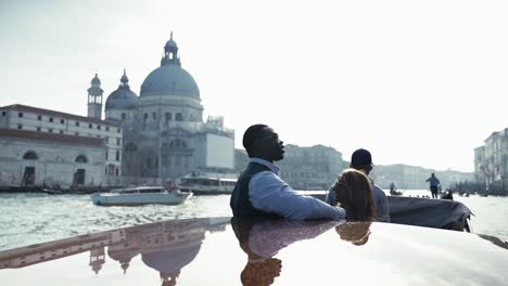 CLassy-mixed-race-couple-having-private-sightseeing-boat-tour-on-the-grand-canal-in-front-of-Basilica-di-Santa-Maria-della-Salute-traveling-Venice-Italy-with-traditional-gondolas-in-scenic-background