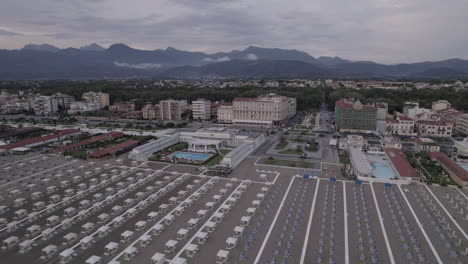 Drone-shot-above-the-beaches-in-Viareggio-Italy-during-sunset-on-a-grey-day-flying-towards-the-buildings-and-hotels-with-mountains-in-the-background-LOG