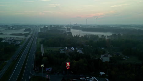 Aerial-drone-top-down-shot-over-cars-passing-over-an-expressway,-passing-though-a-countryside-with-windmills-visible-in-the-background-on-a-foggy-morning