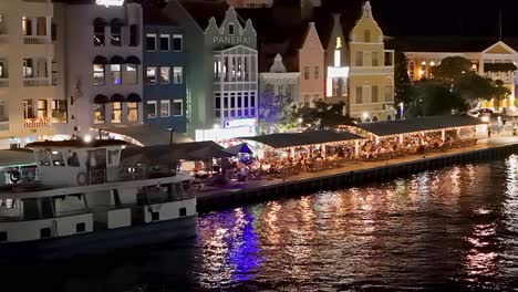 Nachts-Flackern-Die-Lichter-Hinter-Der-Fähre,-Während-Die-Lichter-Auf-Der-Wasserpromenade-Von-Handelskade-Curacao-Schimmern,-Während-Touristen-Mit-Blick-Auf-Das-Meer-Essen