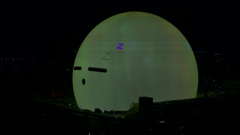 Illuminated-MSG-Sphere-With-Sleeping-Emoji-Face-And-Waking-Up-On-Surface-Looking-Around-At-Night