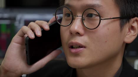 Young-Asian-businessman-in-glasses-engaged-in-a-phone-conversation-with-blurred-stock-market-screens-in-background,-close-up