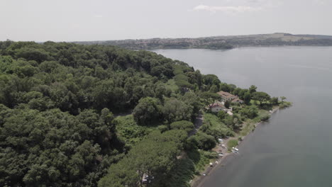 Drone-shot-flying-near-the-edge-of-the-trees-at-Lago-di-Bracciano-Italy-on-a-bright-and-sunny-day-with-cars-driving-on-the-road-near-the-coastline-of-the-lake-LOG