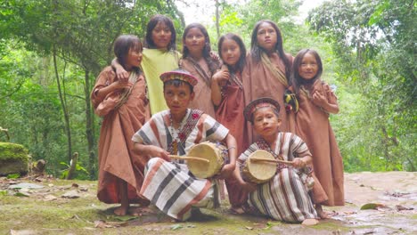 Indigenous-children-in-traditional-attire-playing-drums-and-waving-in-Pucallpa,-Peru,-surrounded-by-lush-forest