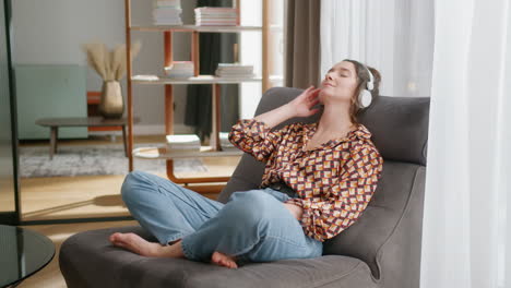 Young-woman-with-headphones-relaxes-in-living-room-with-eyes-shut,-pan