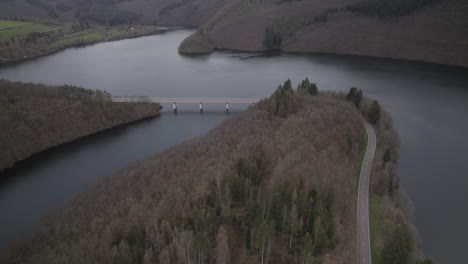 Drone-shot-in-Luxemburg-near-the-river-Lac-de-la-Haute-Sure-at-a-nature-park-Öewersauer-on-a-cloudy-day-with-cars-driving-on-the-road-near-the-forest-and-trees-and-a-bridge-in-the-background-LOG