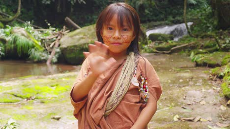 Young-indigenous-girl-waving-in-Pucallpa,-Peru,-with-lush-forest-and-waterfall-background,-daylight