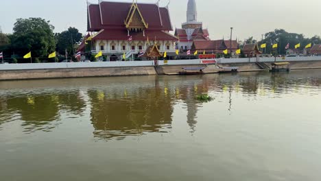 Goldenes-Sonnenlicht-Spiegelt-Sich-Auf-Dem-Fluss-Neben-Einem-Thailändischen-Tempel-Mit-Traditioneller-Architektur-Und-Angedockten-Booten