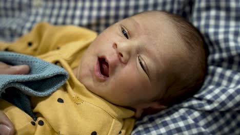 Newborn-baby-boy-in-yellow-onesie-yawning-or-crying-on-checkered-blanket,-close-up
