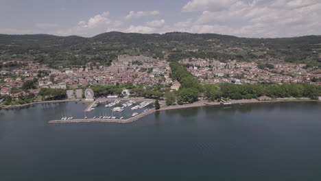 Drone-shot-flying-over-Lake-Bolsena-in-Italy-coming-from-the-water-into-the-old-medieval-city-with-a-castle-and-old-buildings-on-the-hill-and-a-small-harbour-on-a-sunny-day-LOG