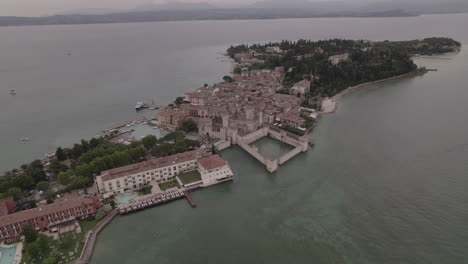 Toma-De-Drone-De-Simione-Italia-Mirando-La-Antigua-Fortaleza-En-Un-Día-Gris-Con-Barcos-En-El-Agua-Cerca-Del-Lago