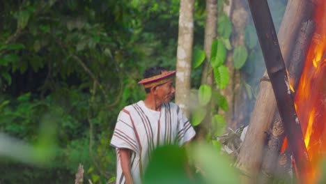 Man-in-traditional-clothing-by-a-fire-in-the-Peruvian-jungle,-leaves-in-foreground,-blurred