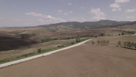 Drohnenaufnahme-Von-Kurvenreichen-Straßen-Und-Goldenen-Feldern-In-Der-Landschaft-Der-Toskana,-Italien,-An-Einem-Sonnigen-Tag-Mit-Blauem-Himmel-Und-Wolken-Am-Horizont