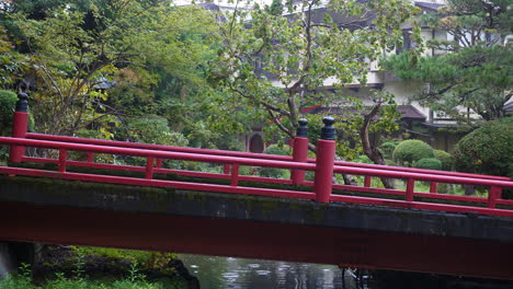 Red-bridge-over-tranquil-pond-in-Koyasan-garden,-surrounded-by-lush-greenery,-serene-atmosphere