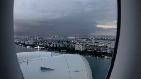 From-the-Airplane-Window,-Witnessing-City-Buildings-Along-the-Singapore-Coastline-Illuminated-by-Lightning-Amidst-Dark-Clouds---POV
