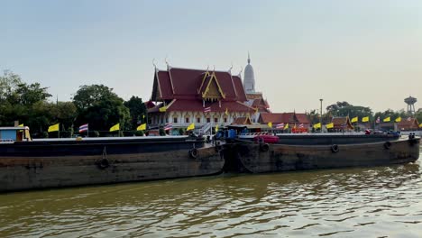 Traditional-Thai-temple-by-river,-barge-in-foreground,-clear-sky,-late-afternoon,-cultural-landscape
