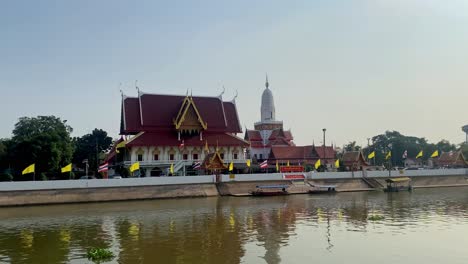 Traditional-Thai-temple-by-river-with-boats,-flags-fluttering,-clear-sky,-serene-setting,-daytime-shot,-wide-angle