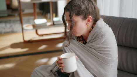 Woman-with-cold-sits-wrapped-in-blanket-and-drinks-tea-in-living-room